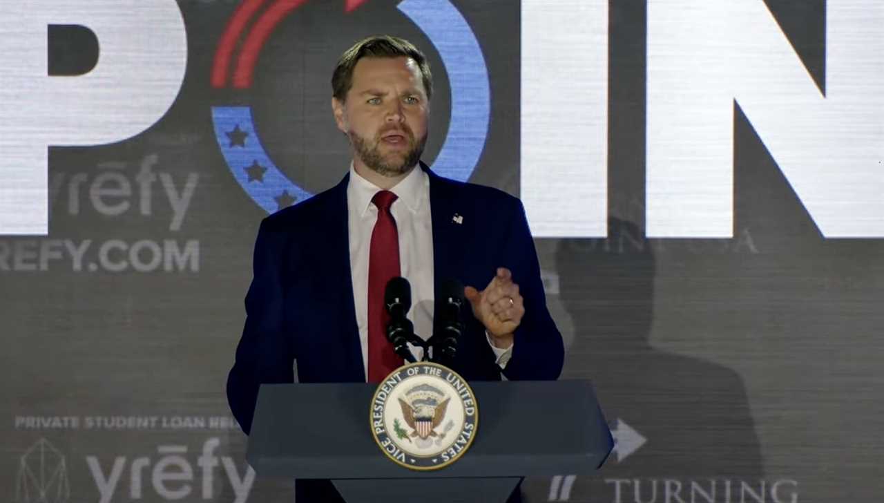 Speaker at a podium with the Vice President seal, addressing an audience during a political event, with a large backdrop displaying the word 