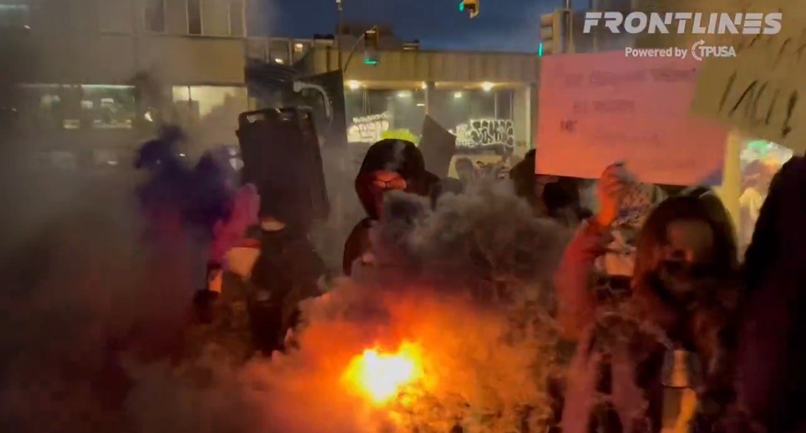 Protesters gather in a smoky urban environment, holding signs and expressing their views during a nighttime demonstration.