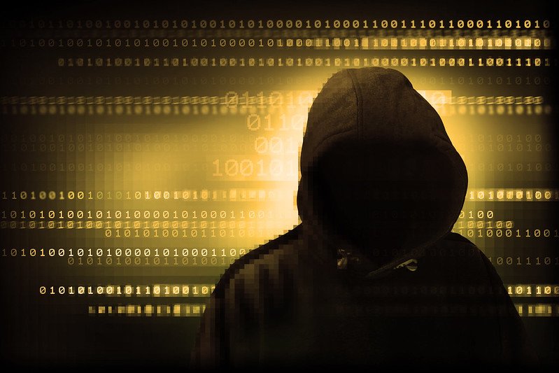 Silhouetted figure in a hoodie against a backdrop of binary code, symbolizing cybersecurity and digital anonymity.
