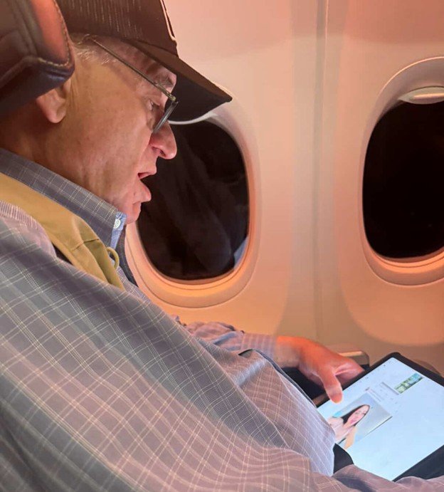Person using a tablet while seated on an airplane, looking at a profile picture, with airplane windows in the background.