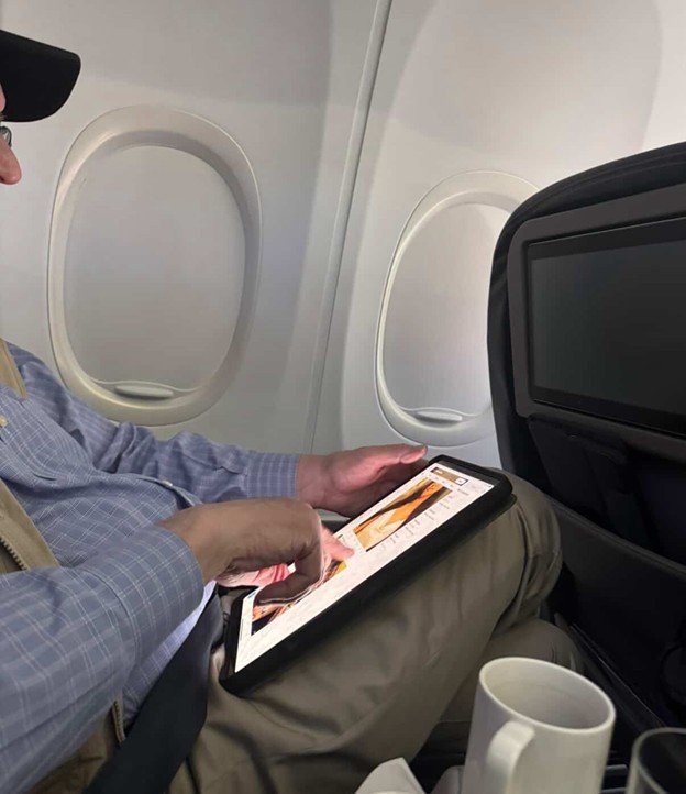 Passenger using a tablet on an airplane, seated comfortably with a coffee cup nearby, showcasing modern travel convenience.