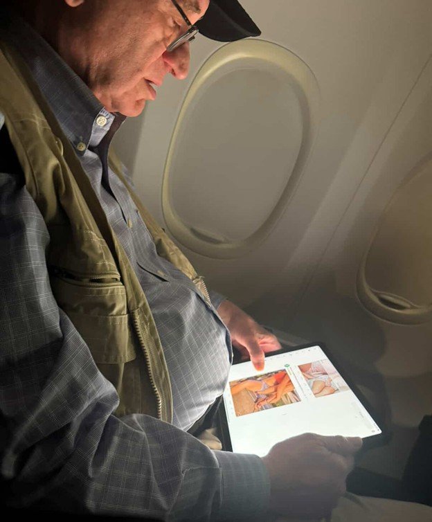 A man reading on a tablet while sitting on an airplane, with airplane windows visible in the background.