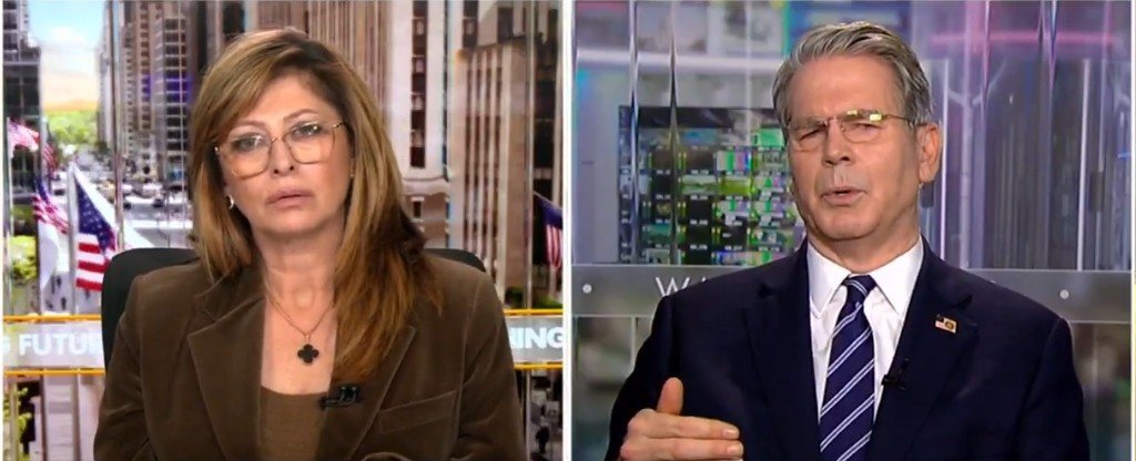 Two news anchors discussing economic topics on a television broadcast, set against a backdrop of city buildings and American flags.