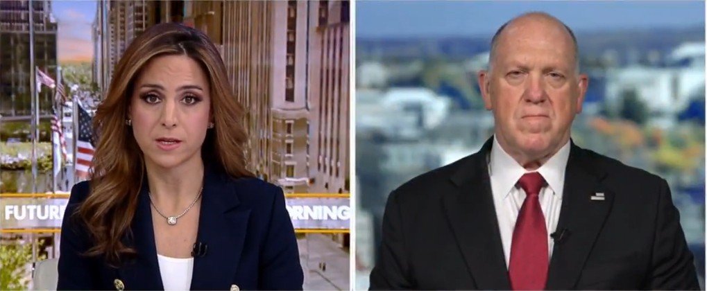 News anchor interviews a guest on a morning show set, discussing current events with a cityscape backdrop featuring American flags.