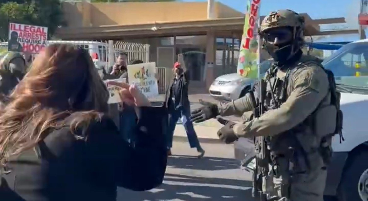 Protesters confront a heavily armed officer during a demonstration against illegal arrests, capturing a tense moment in a public space.