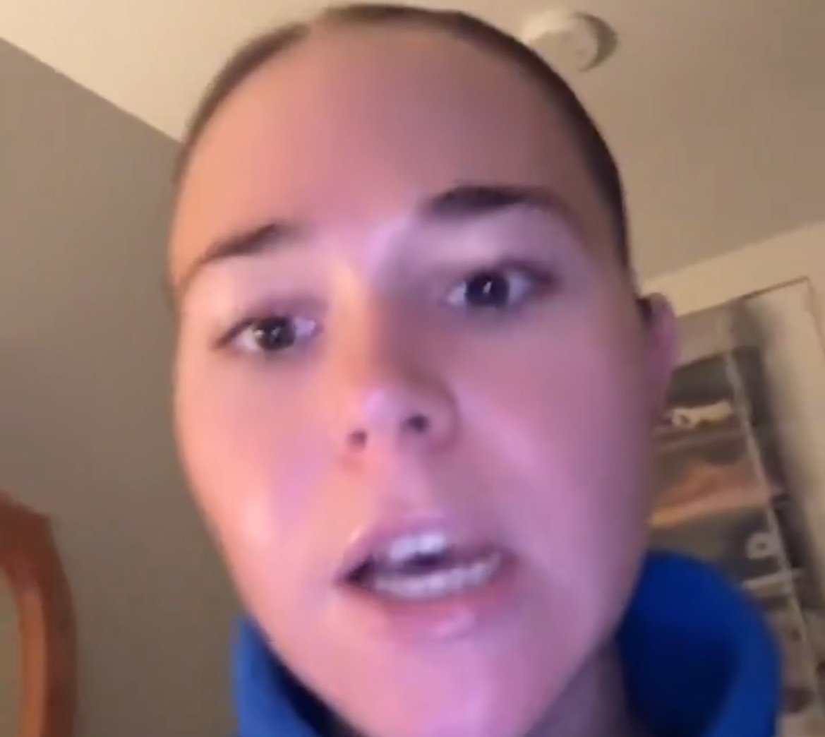 Young woman speaking directly to the camera in a casual setting, wearing a blue hoodie, with a focused expression and indoor background.