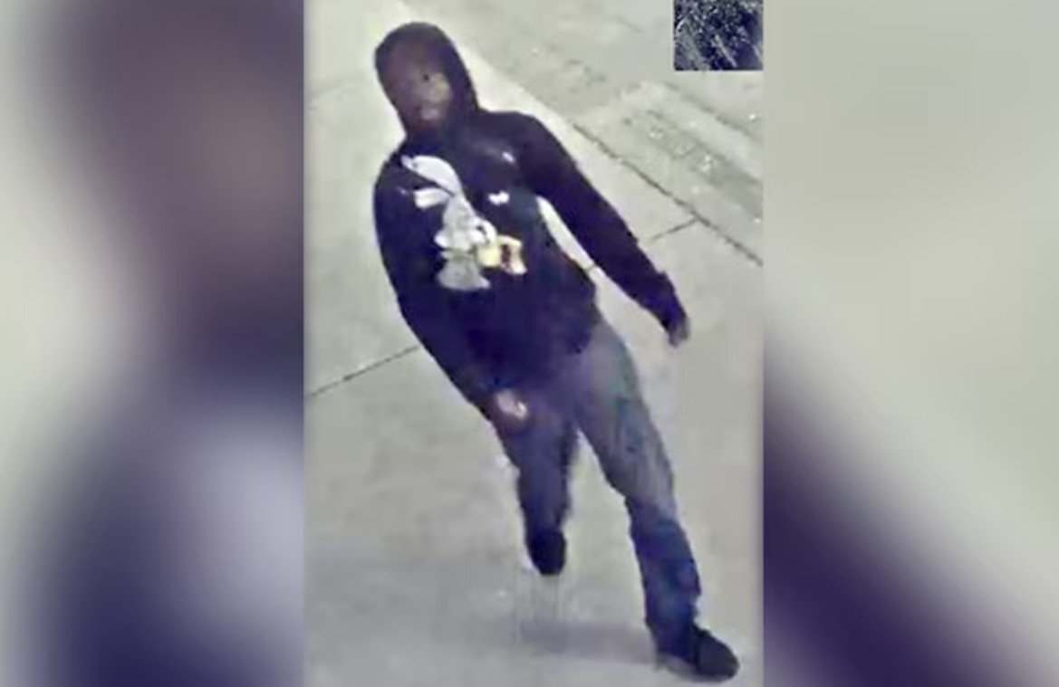 Surveillance image of a person wearing a black hoodie with a graphic, walking on a sidewalk in a city setting.