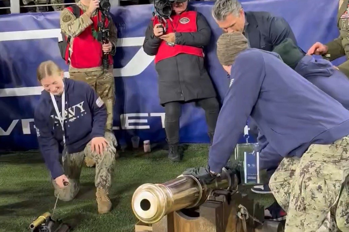 Navy personnel prepare to fire a ceremonial cannon at a sporting event, with onlookers capturing the moment with cameras.
