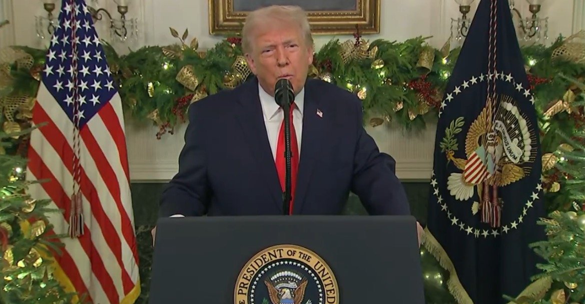 Donald Trump delivering a speech at the White House, decorated for the holidays, with the U.S. flag and presidential seal in the background.