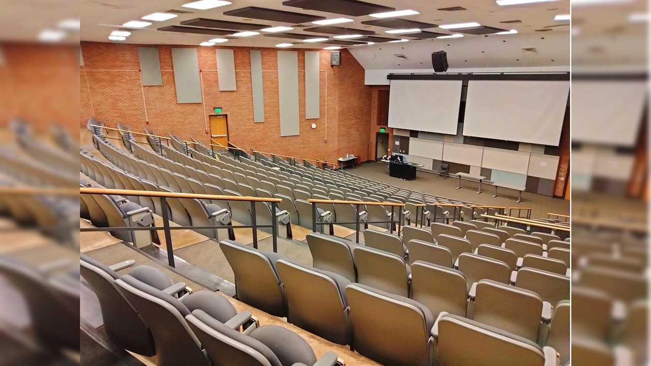 A spacious lecture hall featuring tiered seating, large projection screens, and a central teaching area, suitable for educational presentations and classes.