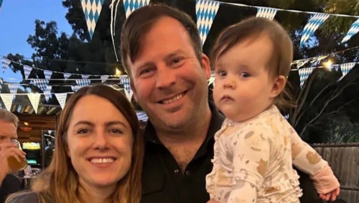 Happy family portrait featuring a couple with their baby, set against a festive outdoor backdrop with decorative bunting.