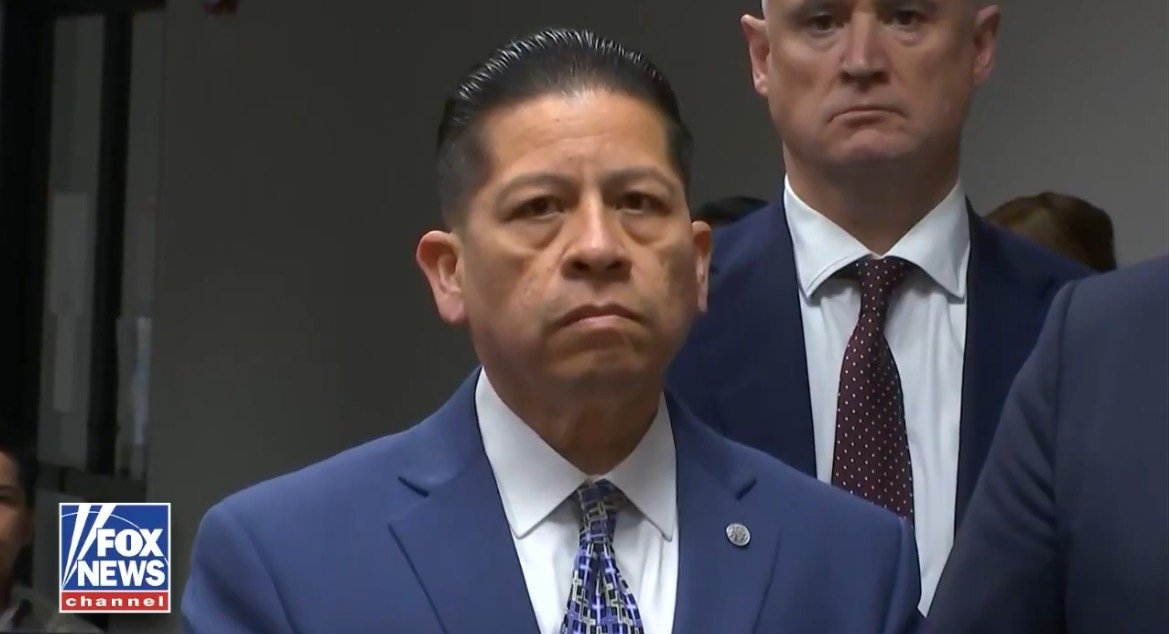 A serious-looking man in a blue suit stands in front of a group of people, conveying a sense of authority during a press event.