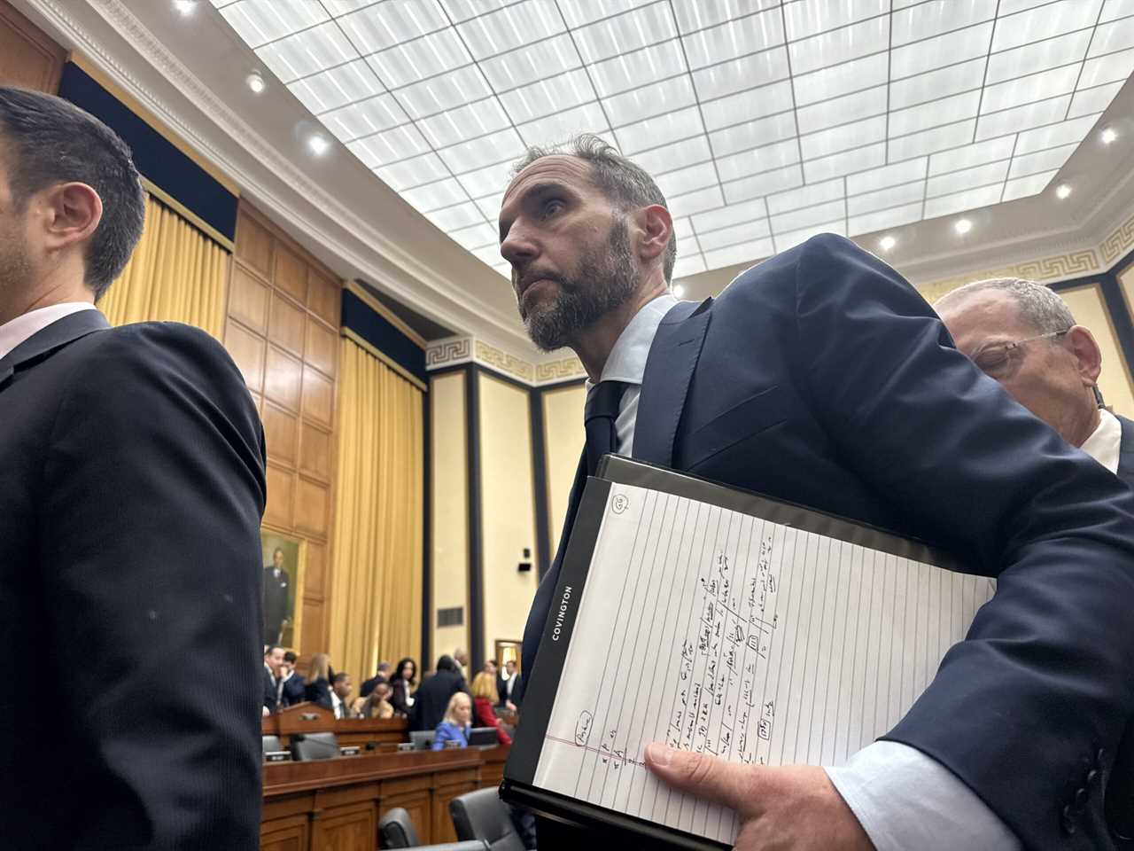 A man in a suit holds a notebook while walking through a congressional hearing room filled with people and wooden furniture.