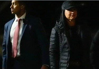 Two individuals walking side by side at night, one in a suit and tie, the other in a black jacket and cap.