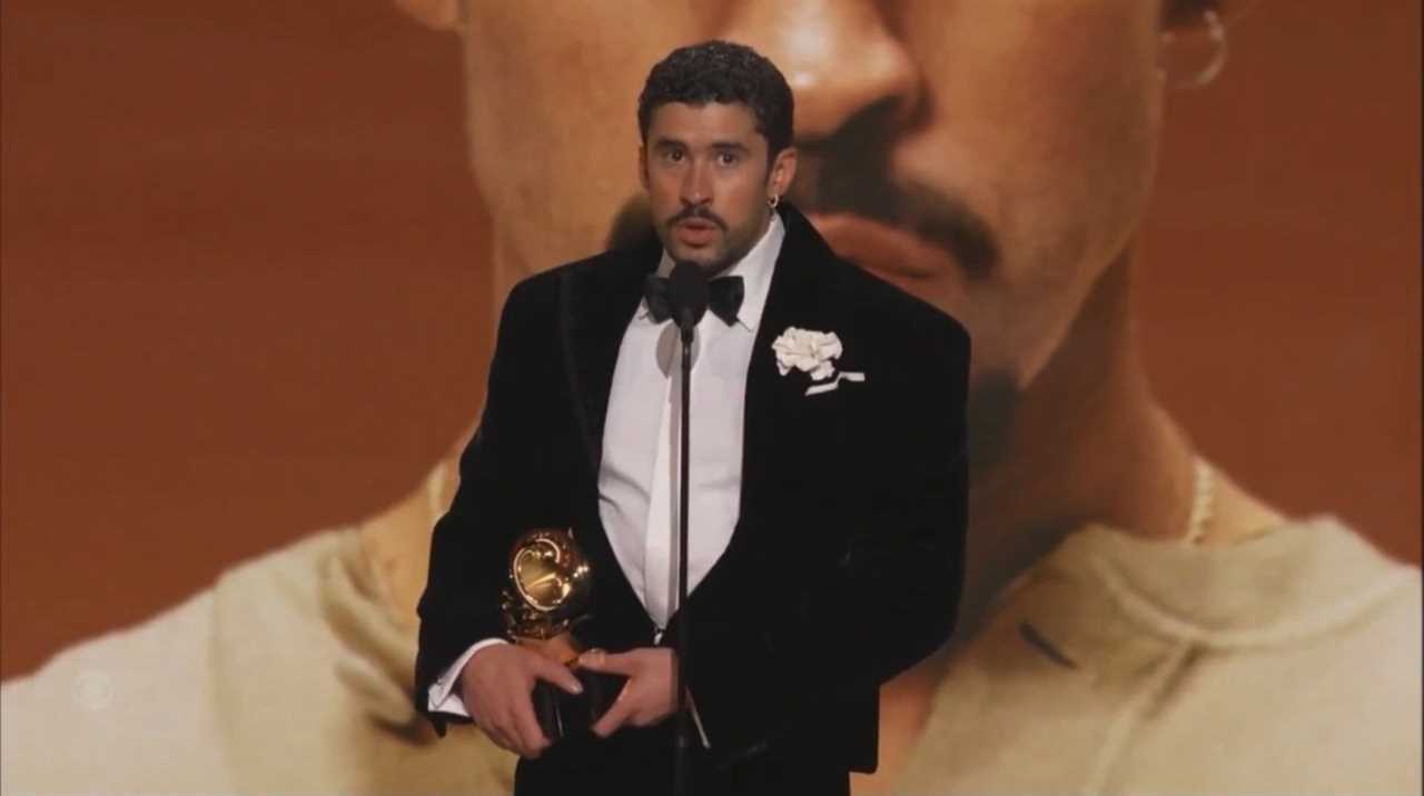 Award recipient delivering a speech at a prestigious ceremony, holding a trophy, with a large artistic backdrop.