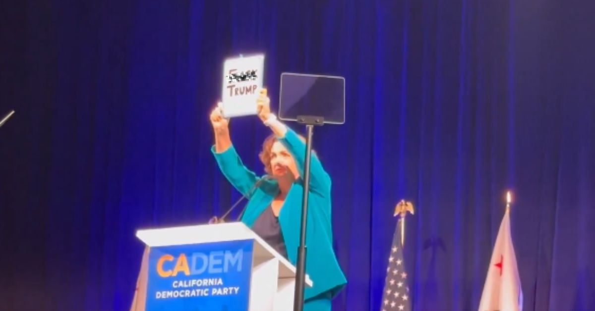 Speaker at California Democratic Party event holds a sign expressing opposition to Trump, with flags and podium in the background.