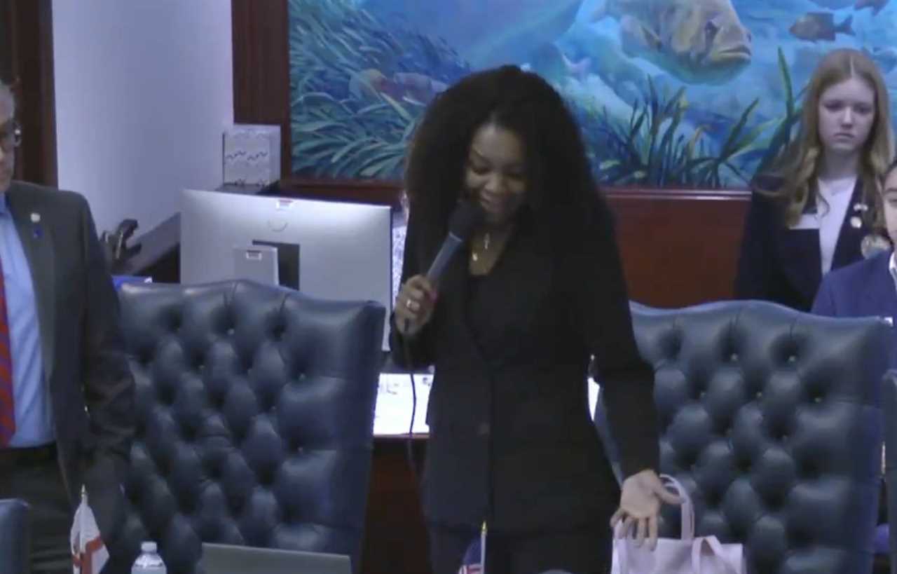 A woman speaks into a microphone in a legislative setting, with colleagues and a colorful mural in the background, highlighting civic engagement and dialogue.