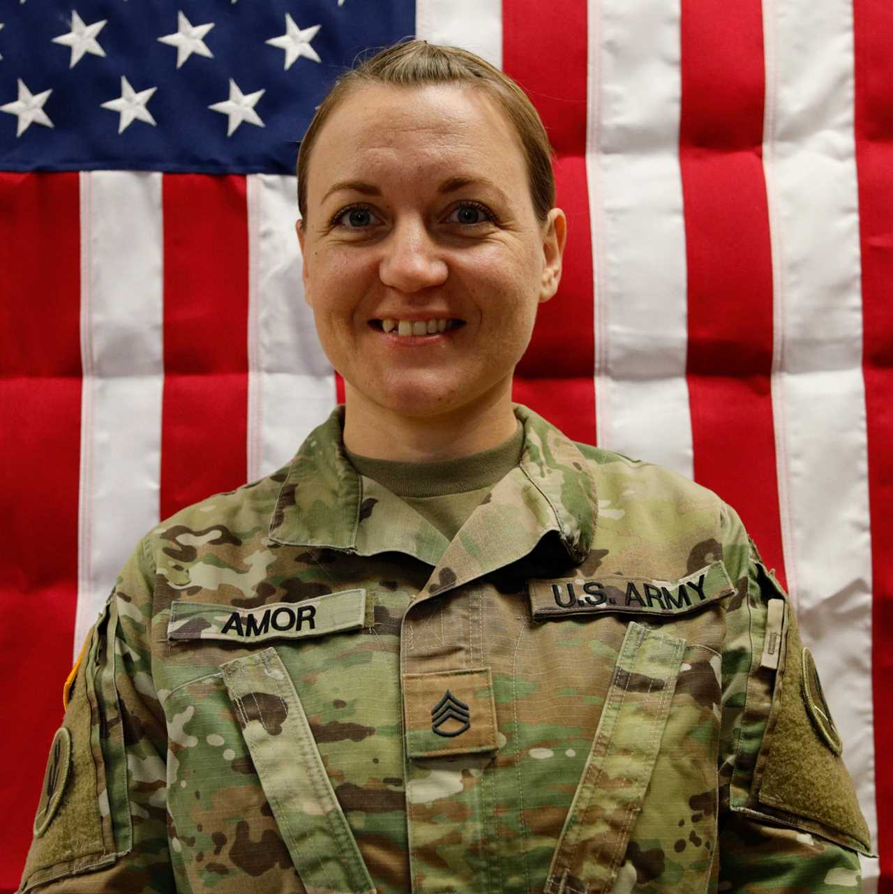 U.S. Army soldier in uniform smiles in front of an American flag backdrop, showcasing pride and dedication to service.