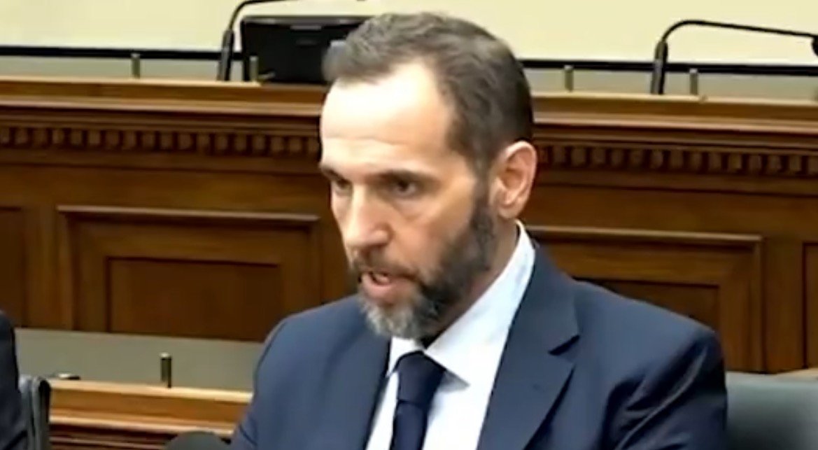 Man in a suit speaking during a formal meeting, with a wooden backdrop and empty seats visible, conveying a serious tone.