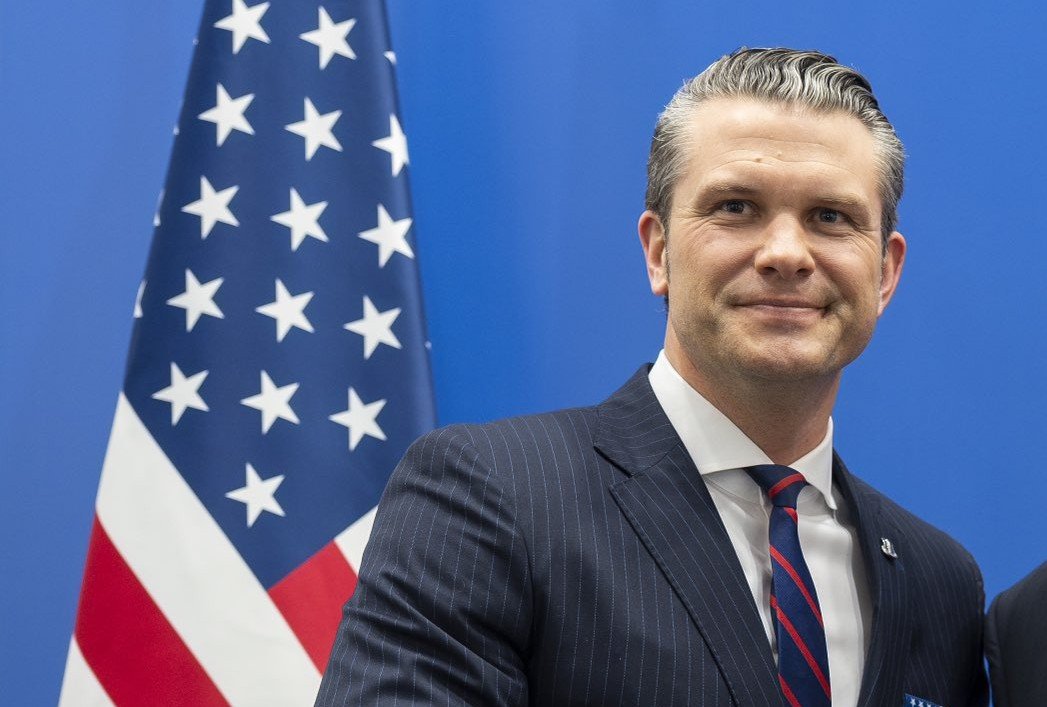 Man in a pinstripe suit stands beside the American flag, smiling against a blue background during an official event.