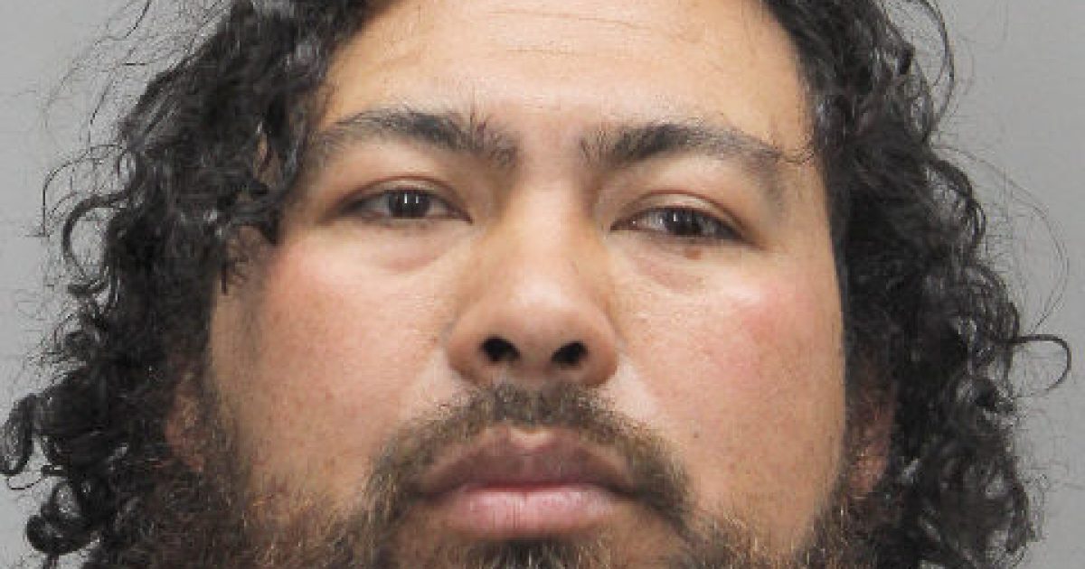 Mugshot of a man with curly black hair and a beard, wearing a white jumpsuit, against a gray background.