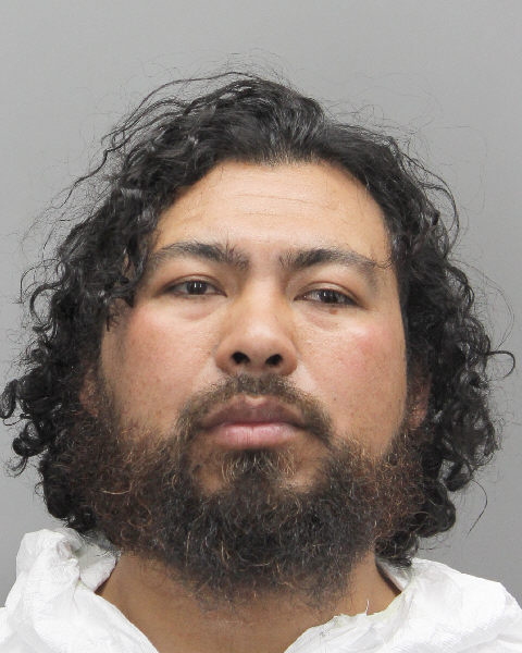 Mugshot of a man with curly black hair and a beard, wearing a white jumpsuit, against a gray background.