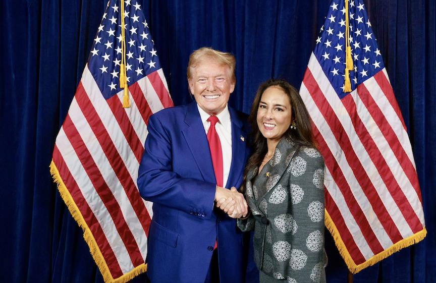Donald Trump and a woman shaking hands in front of two American flags, smiling at the camera during a formal event.