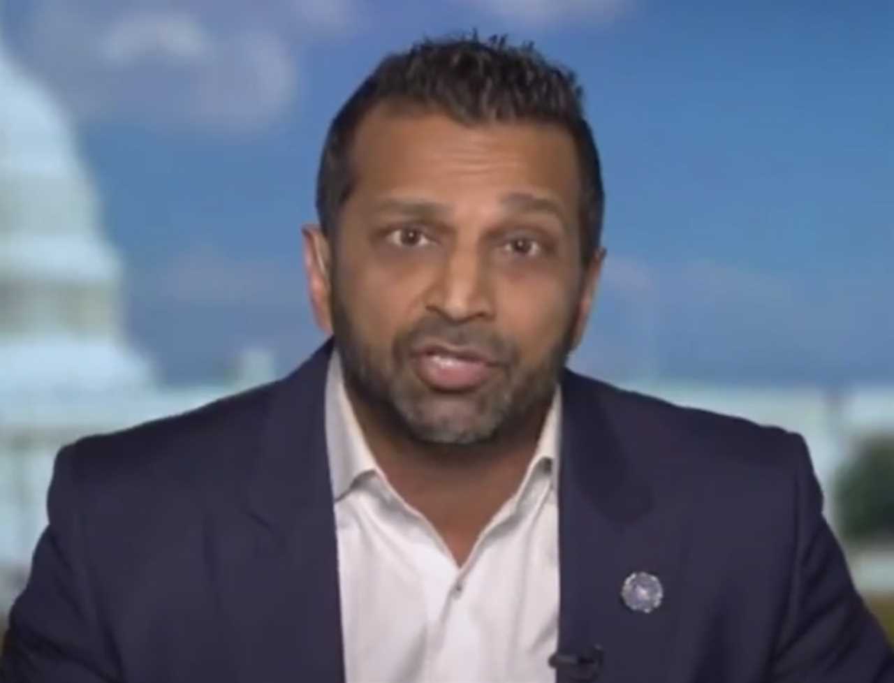 Man speaking on a news program with a blurred background of a government building, wearing a dark suit and white shirt.