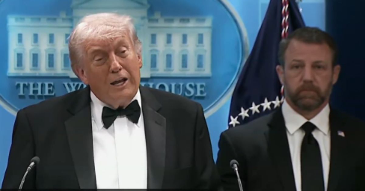 Donald Trump speaks at a press conference in the White House, wearing a tuxedo, with a security detail standing beside him.