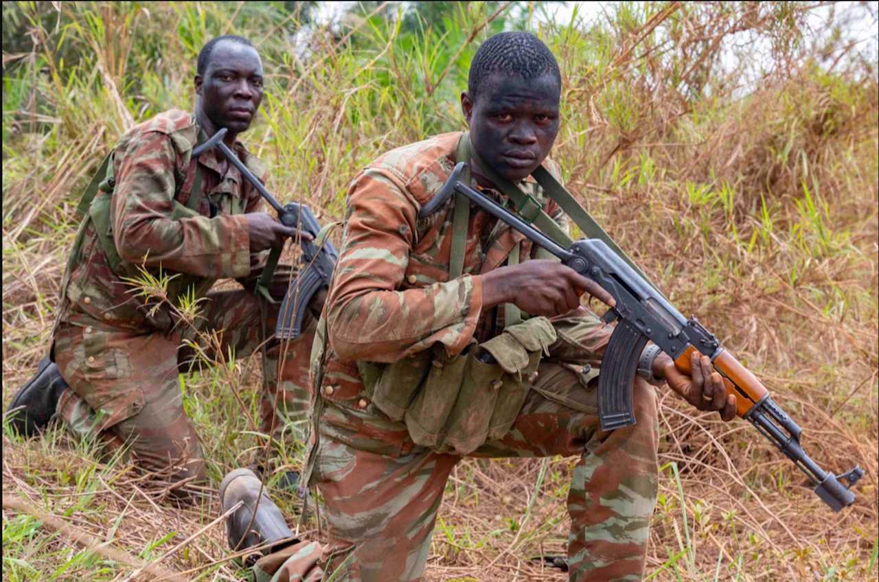 Two soldiers in camouflage uniforms crouch in tall grass, holding rifles as they prepare for action in a military training or operational setting.