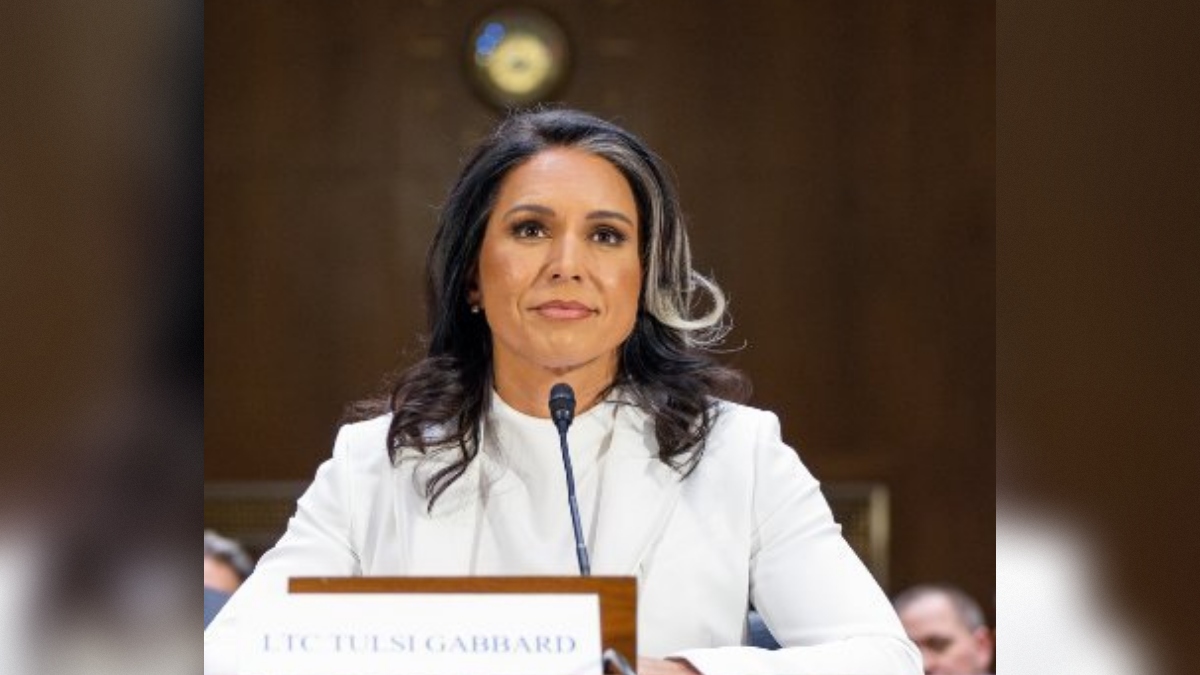LTC Tulsi Gabbard testifies during a congressional hearing, seated at a table with a microphone, dressed in a white blazer, showcasing a serious expression.