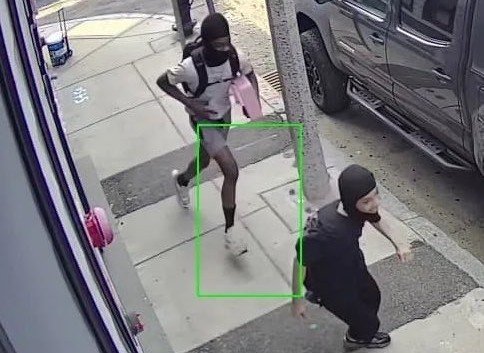 Two individuals in casual attire walking on a sidewalk, one wearing a backpack and the other in dark clothing, captured by surveillance camera.