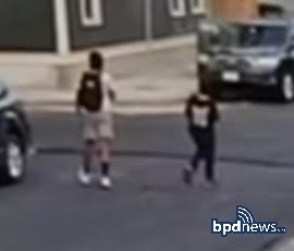 Two individuals walking on a street near parked cars, with one carrying a backpack and both dressed casually.