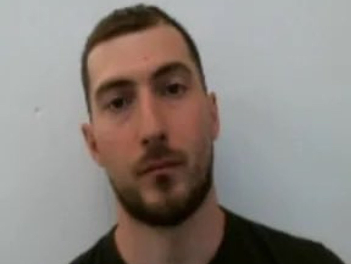 Man with short brown hair and a beard, wearing a black shirt, posing against a plain white background.