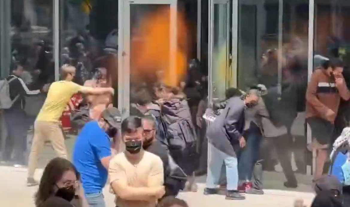 Crowd of people gathered outside a glass building, some attempting to enter while others observe, during a chaotic scene.