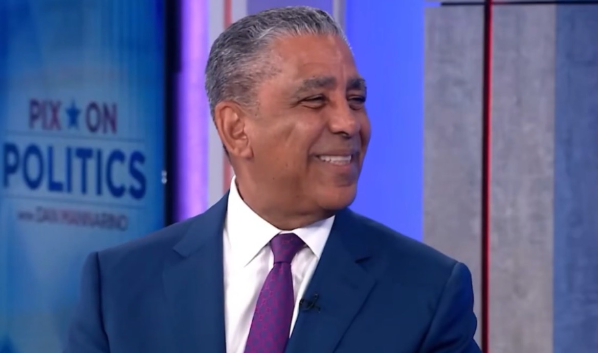 Smiling man in a blue suit with a purple tie, seated in a studio setting, discussing politics on a television show.