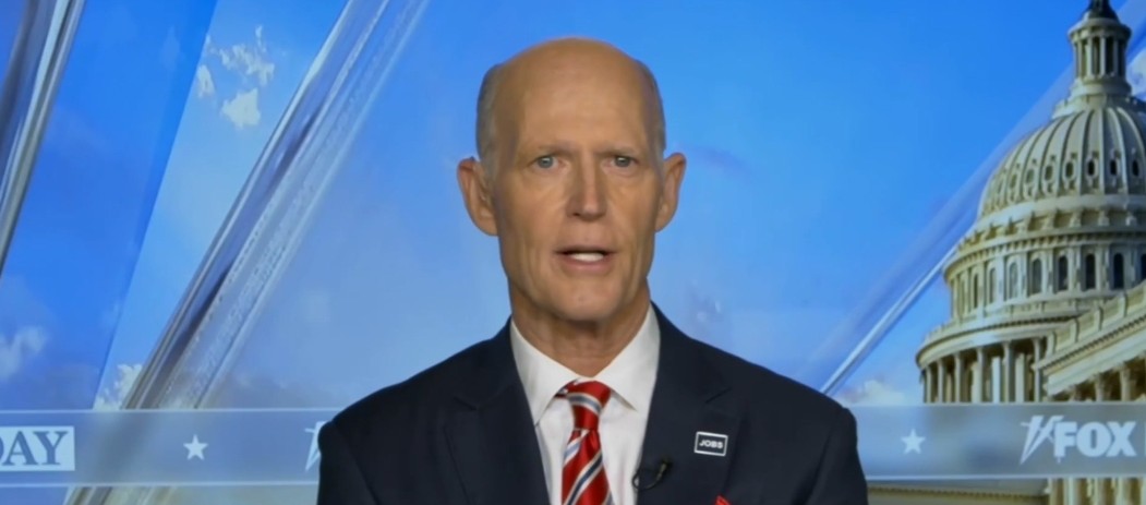 Rick Scott speaking during a televised interview, with the U.S. Capitol building visible in the background, highlighting political themes and discussions.