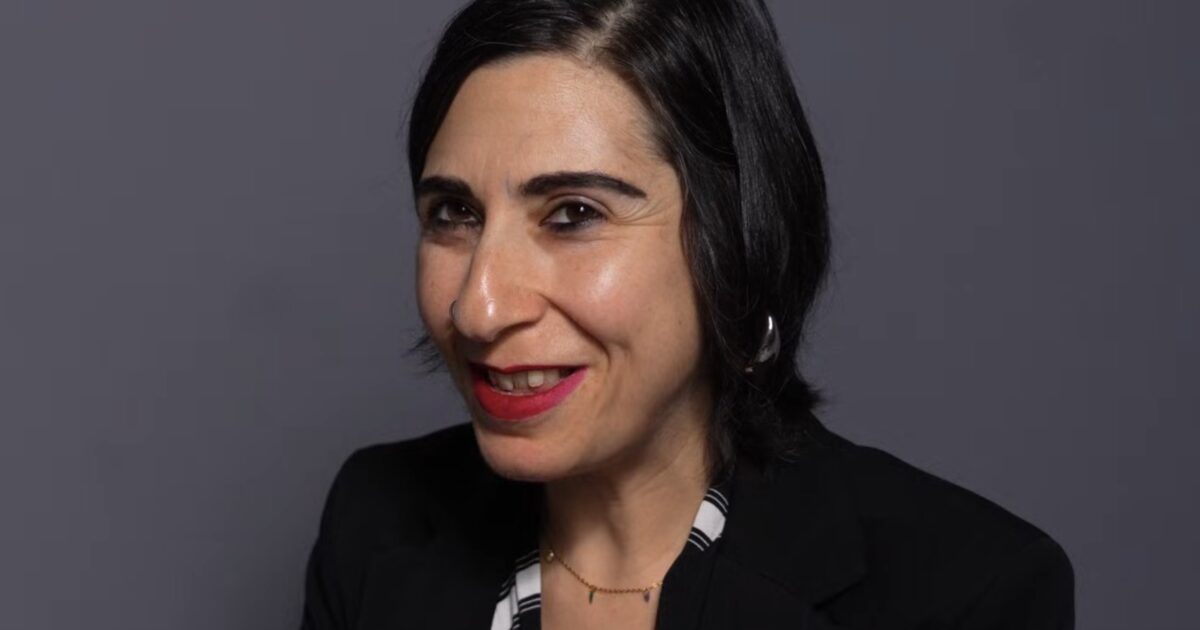 Portrait of a smiling woman with dark hair and red lipstick, wearing a black blazer against a gray background.