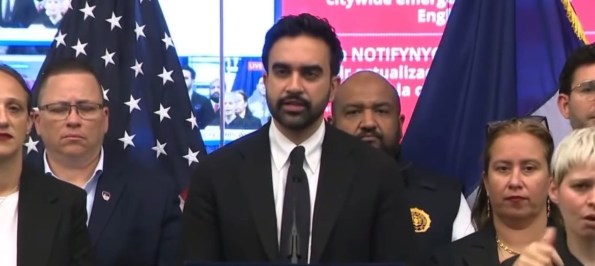 Press conference with a speaker addressing the audience, flanked by officials and an American flag, discussing important community issues.