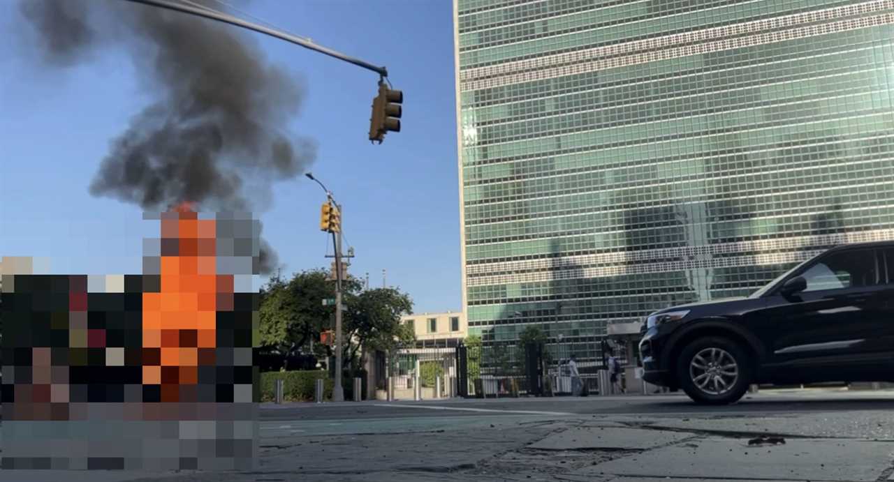 Fire and smoke billow from a vehicle on the street near a modern building, with traffic signals visible in the foreground.