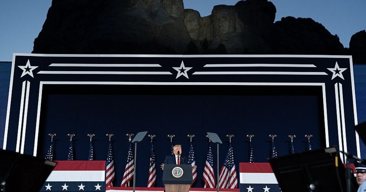 Donald Trump delivers a speech at a patriotic event with American flags and a dramatic mountain backdrop during twilight.