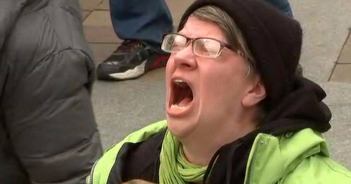 Person in a green jacket and black beanie expressing strong emotions while sitting on the ground during a protest.