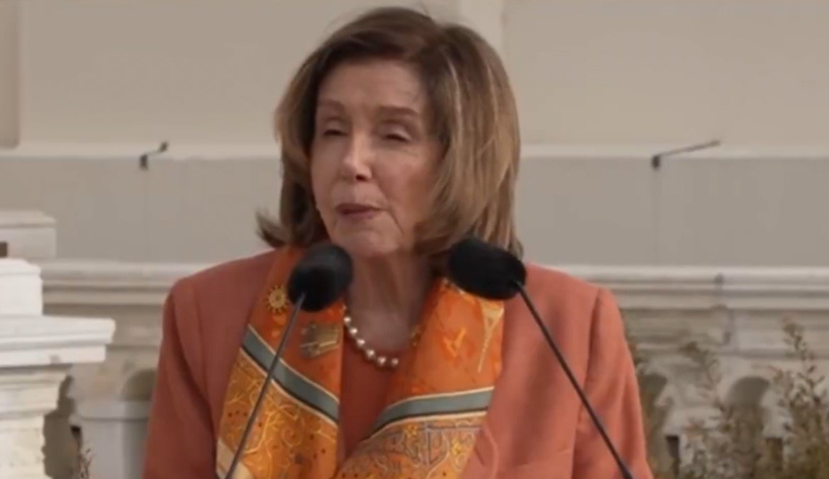 Speaker delivering a speech outdoors, wearing an orange blazer and a decorative scarf, with microphones positioned in front of her.